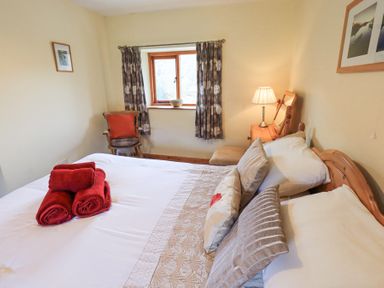 A bedroom with a double bed with pillows and red towels on it a wooden chair with a cushion a window with curtains and a wooden dresser with a lamp at Low Wood View in Hartsop