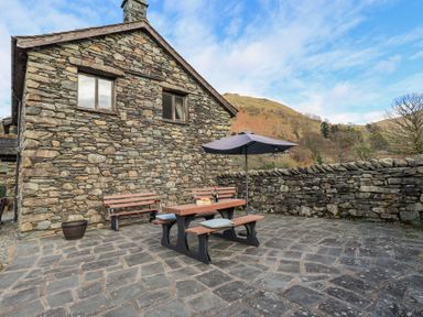 An outdoor stone patio with a picnic table umbrella and bench against a stone house and wall at Low Wood View in Hartsop