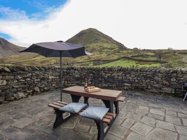 An outdoor patio with a picnic table cushions umbrella and a stone wall with hills in the background at Low Wood View in Hartsop