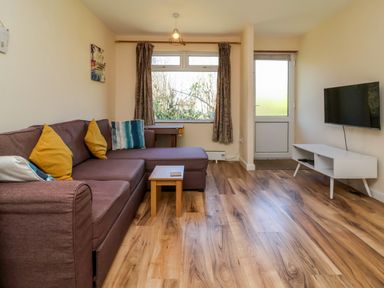 A living room with a brown sectional sofa and yellow pillows a small wooden table a wall mounted television and a window at 24 The Glade in Kilkhampton