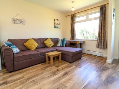A living room with a brown sectional sofa with cushions a small wooden table wooden flooring and a window with curtains at 24 The Glade in Kilkhampton