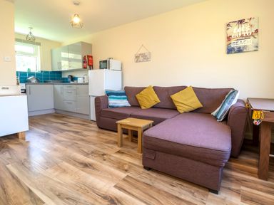 A living room with a brown sofa and cushions next to a small wooden table and an open kitchen area with grey cabinets and a white refrigerator at 24 The Glade in Kilkhampton