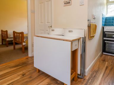 A kitchen area with a foldable dining table, two white chairs, a stove, a towel hanging on a wall rack, and a view of a bedroom with two wooden beds at 24 The Glade in Kilkhampton