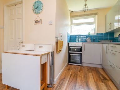 A kitchen with white cabinets blue tiled backsplash a small oven a table with two white chairs a towel hanging on the wall and wooden flooring at 24 The Glade in Kilkhampton