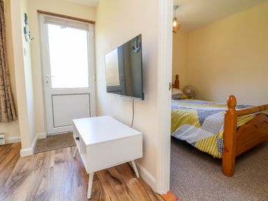 A small hallway with a mounted TV and white media unit next to a door and a bedroom with a wooden bed at 24 The Glade in Kilkhampton