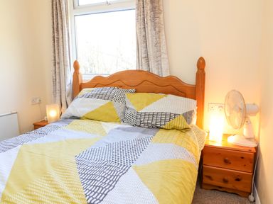 A bedroom with a wooden bed and yellow patterned bedding with curtains on the window at 24 The Glade in Kilkhampton