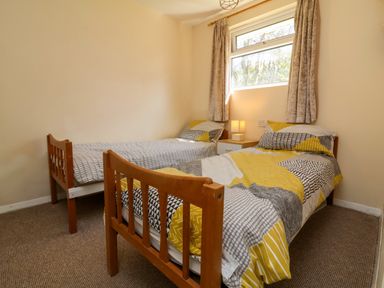 A bedroom with two single wooden beds a nightstand with a lamp and a window with curtains at 24 The Glade in Kilkhampton