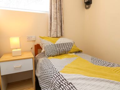 A single bed with patterned bedding next to a wooden nightstand with a lamp at 24 The Glade in Kilkhampton