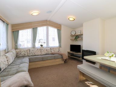 A living room with an L-shaped sofa, a TV on a wooden stand, shelves with decorations, and a dining table with a bench at 31B Foxburrow Hang in Fritton near Belton Norfolk