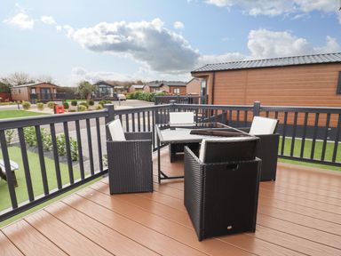 An outdoor patio with black wicker chairs with white cushions and a glass table surrounded by wooden buildings at Blossom Vale Lodge in Evesham