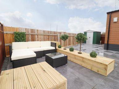 An outdoor patio with a sectional sofa white cushions glass table wooden seating planters and a green shed in the background at Blossom Vale Lodge in Evesham