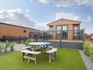A yard with a round wooden picnic table on artificial grass in front of a small wooden lodge with a deck and railing at Blossom Vale Lodge in Evesham
