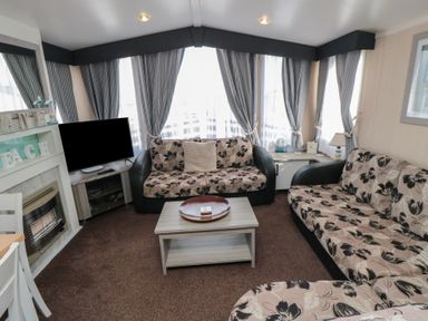 A living room with two floral sofas a TV a fireplace a coffee table and large windows with curtains at Rose Bank D8 in Primrose Valley