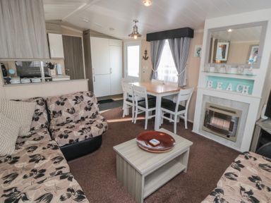 A living room with a floral sofa, wooden coffee table, dining table with chairs, and a fireplace at Rose Bank D8 in Primrose Valley