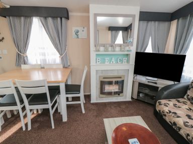 A dining table with chairs next to a fireplace with decorative beach letters and a television in a living room at Rose Bank D8 in Primrose Valley