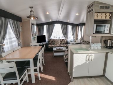 A dining area with a wooden table and white chairs next to a living room with sofas and a television at Rose Bank D8 in Primrose Valley