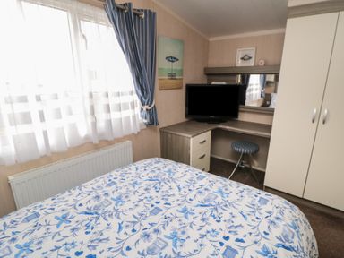 A bedroom with a bed with blue and white floral bedding a window with curtains a desk with a television a round stool and a wardrobe at Rose Bank D8 in Primrose Valley