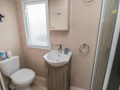 A bathroom with a toilet, sink with cabinet, window with blinds, and a shower at Rose Bank D8 in Primrose Valley