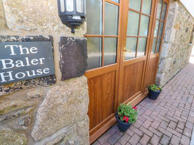 A stone wall with a sign reading The Baler House next to wooden doors with glass panels and pots with flowers on the ground at The Baler House in Hayle