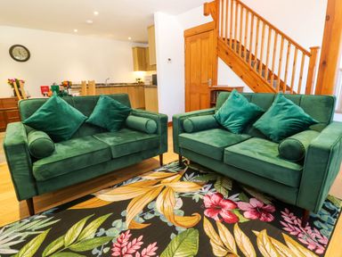 A living area with two green sofas on a floral rug near a wooden staircase and kitchen at The Baler House in Hayle