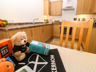 A kitchen with wooden cabinets, a countertop with sink, a wooden chair, and a table with a teddy bear, food boxes, and a black mat at The Baler House in Hayle