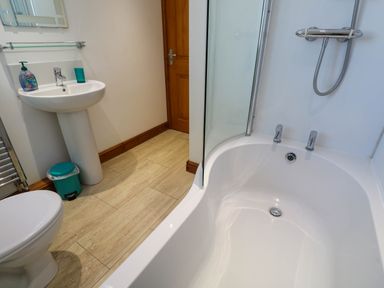 A bathroom with a white bathtub and shower head a white sink with soap and cup a wooden door a toilet and a turquoise trash bin at The Baler House in Hayle