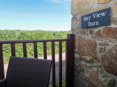 A brown wicker chair and wooden fence next to a stone wall with a Bay View Barn sign with greenery in the background at Bay View Barn in Hayle