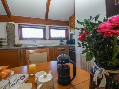 A kitchen with wooden cabinets and two small windows above the sink with a dining table in the foreground holding a French press coffee maker, a teapot, cups, a glass of orange juice, croissants, and a vase with flowers at Bay View Barn in Hayle