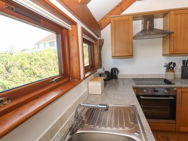 A kitchen with a stainless steel sink an electric stove wooden cabinets and two windows showing greenery at Bay View Barn in Hayle