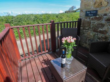 A balcony with two wicker chairs a glass table with a vase of flowers and a bottle of wine at Bay View Barn in Hayle