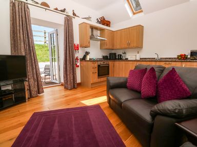 A living area with a black sofa and purple cushions near a kitchen with wooden cabinets and a TV at The Coach House in Hayle