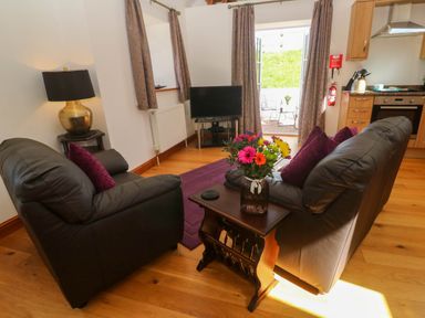 A living room with two black leather sofas a wooden table with flowers a lamp a television and kitchen area at The Coach House in Hayle