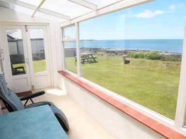 A sunroom with a cushioned chair and table overlooking a lawn with a picnic bench and ocean view at Tegawelon in Llandanwg near Harlech