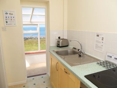 A kitchen with a sink toaster and stovetop with a view of grass and sea through an open door at Tegawelon in Llandanwg near Harlech