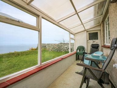 A sunroom with plastic and reclining chairs and large windows showing a grass lawn and a stone wall at Tegawelon in Llandanwg near Harlech