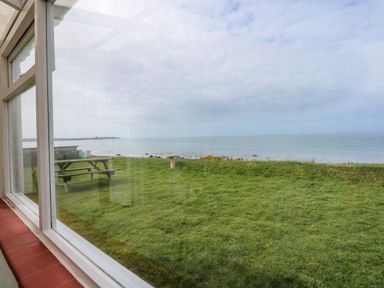 A view of a grassy lawn with a picnic bench and a bird bath by the seaside through a window at Tegawelon in Llandanwg near Harlech