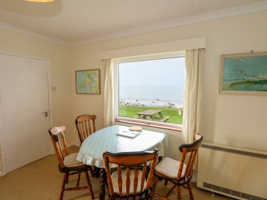 A dining area with a table and four chairs near a window overlooking a picnic table and sea at Tegawelon in Llandanwg near Harlech