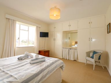 A bedroom with a bed with rolled towels a window a white built in wardrobe a wall mounted tv a wooden bedside table and a wicker chair at Tegawelon in Llandanwg near Harlech