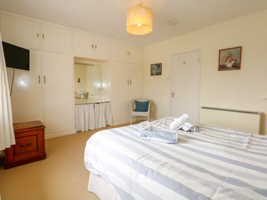 A bedroom with a bed and towels on it a chair a chest of drawers a wall-mounted television and built-in wardrobes with a dressing table at Tegawelon in Llandanwg near Harlech