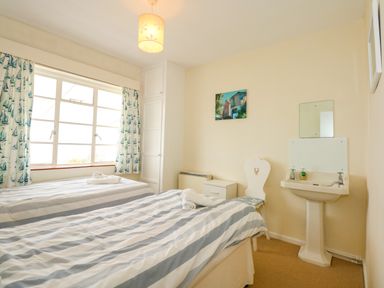 A bedroom with two single beds next to a window with sailboat curtains a white chair a sink and a small cabinet at Tegawelon in Llandanwg near Harlech