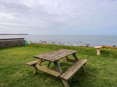 A wooden picnic table on grass near a rocky shore with the sea and cloudy sky in the background at Tegawelon in Llandanwg near Harlech