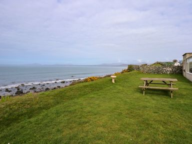 A grassy area with a picnic table and birdbath overlooking a rocky shoreline and sea at Tegawelon in Llandanwg near Harlech