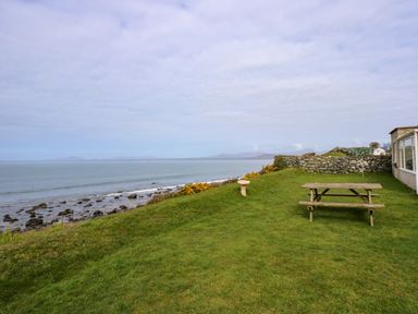 A grassy lawn with a wooden picnic table a birdbath and stone wall by the sea at Tegawelon in Llandanwg near Harlech