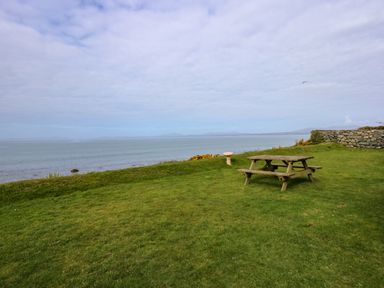 A picnic table and bird bath on a grassy area overlooking the sea with a stone wall on the right at Tegawelon in Llandanwg near Harlech