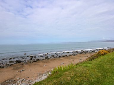 A rocky beach with sand and grass foreground and distant mountains across the water at Tegawelon in Llandanwg near Harlech