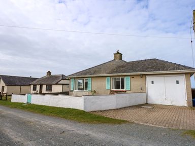A bungalow with turquoise window shutters a white garage door and a low white wall beside a gravel road at Tegawelon in Llandanwg near Harlech