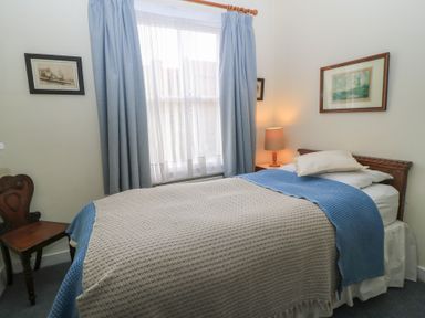 A bedroom with a single bed covered with beige and blue blankets a wooden chair a nightstand with a lamp and framed pictures on walls at The Townhouse in Salisbury