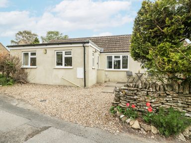 A single-story house with a gravel driveway a stone wall with flower bed and outdoor table and chairs at Spring Cottage in Birdlip near Brockworth