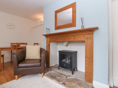 A living room with a brown armchair a wooden dining table and chairs a clock on the wall a fireplace with a wooden mantel and a black stove inside at Spring Cottage in Birdlip near Brockworth