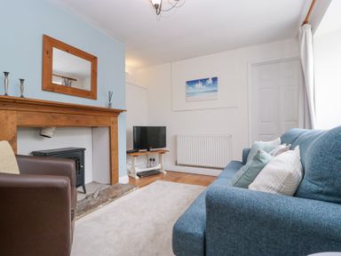 A living room with a blue sofa, a brown armchair, a wooden fireplace mantel, a TV on a wooden stand, and a picture on the wall at Spring Cottage Birdlip near Brockworth